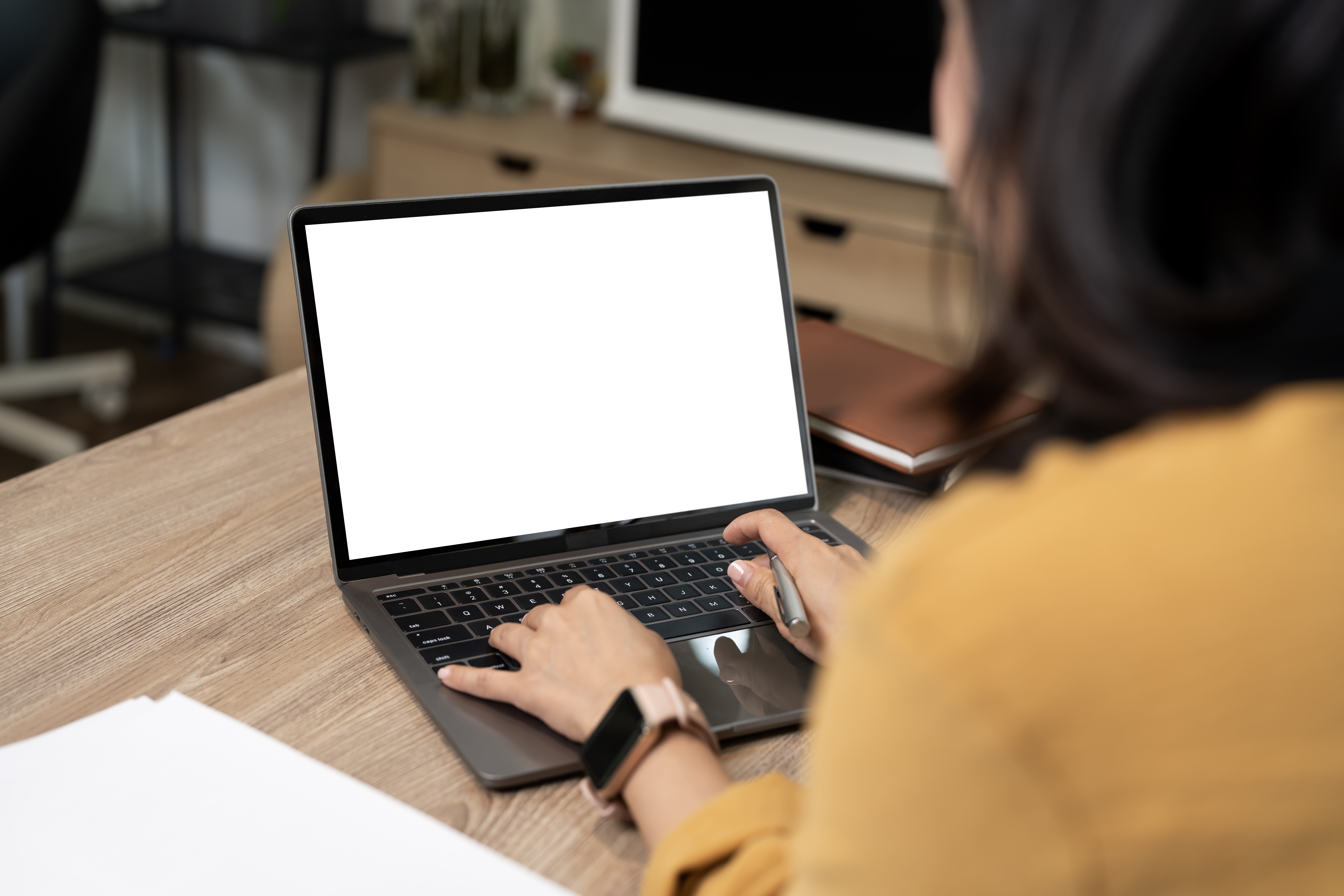 Over the shoulder view of a woman using a laptop with a blank white screen mockup at an office desk.