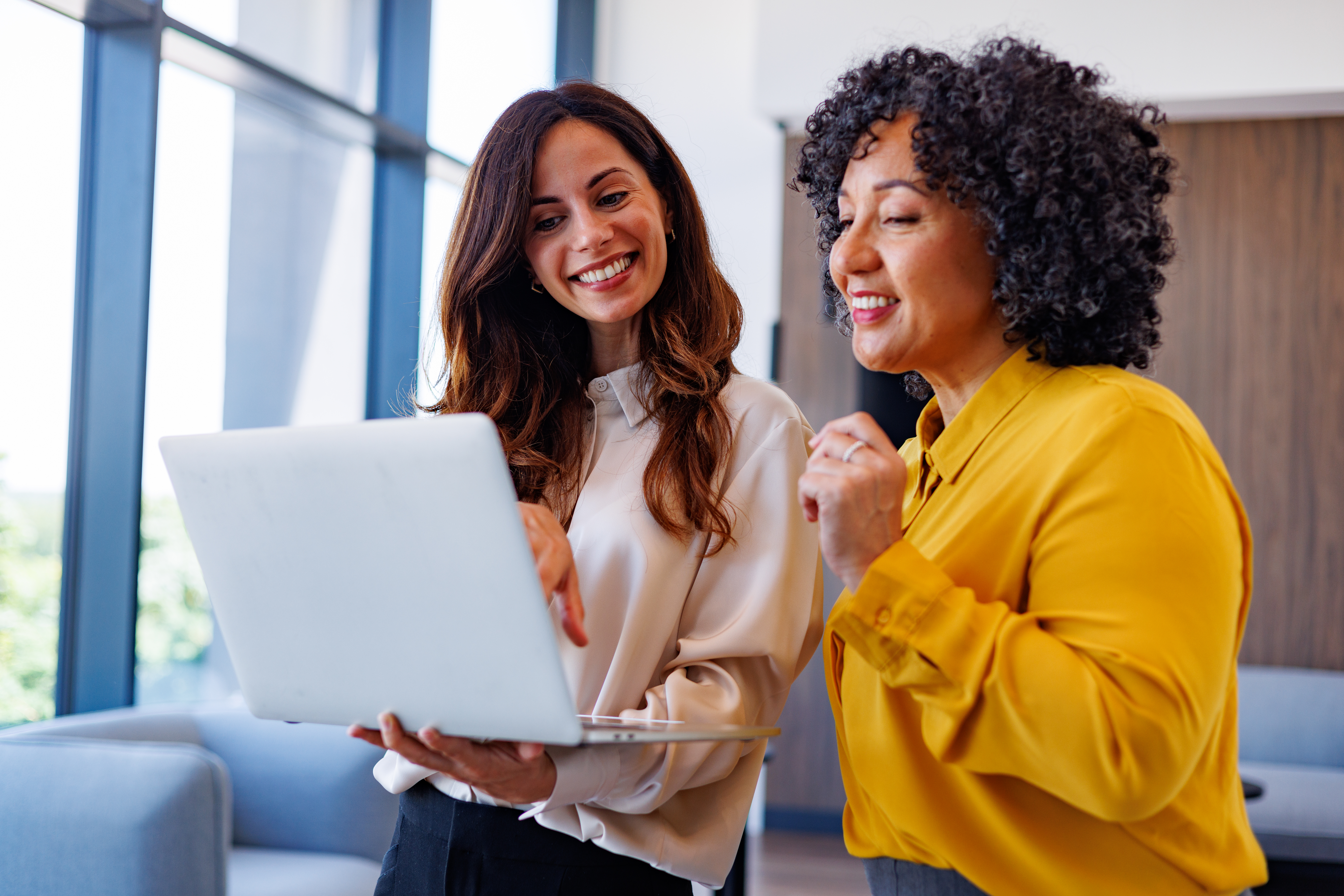 Two smiling businesswomen actively collaborating, looking at a laptop screen and discussing work in a contemporary office setting